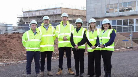 Six people are stood on a construction site. They are all wearing yellow high visibility clothing and white hard hats. The buildings behind them are under construction with large piles of sand in front and scaffolding around them.