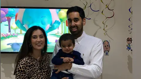 Family handout A couple smile at the camera while the dad holds their young son. The woman has long black hair past her shoulders and wears an animal print top. The man wears a white shirt and has short black hair and a short black beard. The youngster has short black hair and wears a navy top and jeans.