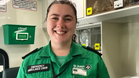 A smiley woman stands in an ambulance She wears a St John Ambulance uniform 