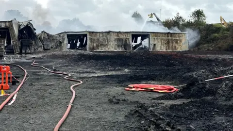 Northamptonshire Fire and Rescue Service A burnt out building, a large fire hose, red plastic barriers on the ground, smoke in the air, and water being poured on a metal building that is crumpled and collapsed