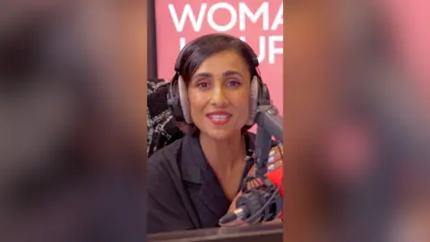 A woman with short, dark hair sits in front of the microphone and speaks to the camera