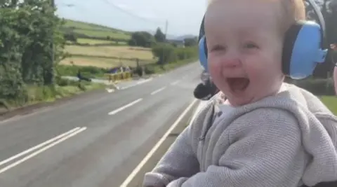 A baby looks at the road with an expression of glee. He wears a grey hooded jumper and blue ear defenders.
