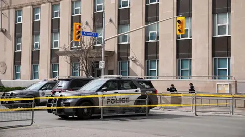 An image taken after the March shooting of the US Consulate building in Toronto. It shows three Toronto police cars behind caution tape and metal fences. Behind them is the building, which is beige and features an entrance and rows of vertical windows. 