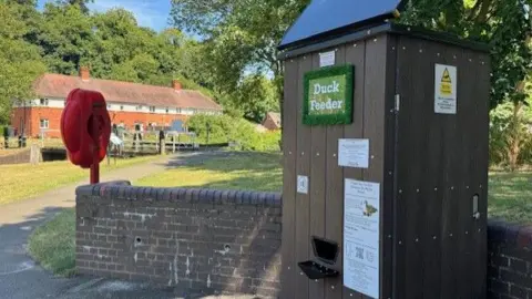 The duck vending machine is pictured at the park next to a small brick wall. It has a solar panel on the roof with a wooden panelled frame. A green sign reads duck feeder. 