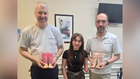 Supplied Olivia Story in black top and smiling at camera. She has long brown hair and a tattoo on her right arm. She is standing between two men who are both wearing grey T-shirts. The man on the left of the photo is holding two pink small prosthetic legs and smiling. He has grey hair. Neil Wallace is on the right holding two white prosthetic legs. On the walls in the background is TV and a piece of wall art. 
