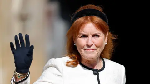 Sarah Ferguson, the then Duchess of York, attends the traditional Easter Sunday Mattins Service at St George's Chapel, Windsor Castle on April 20, 2025. She has long red hair with a black hairband and is dressed in a white jacket with back trim and is wearing black gloves. One arm is raised as she waves.
