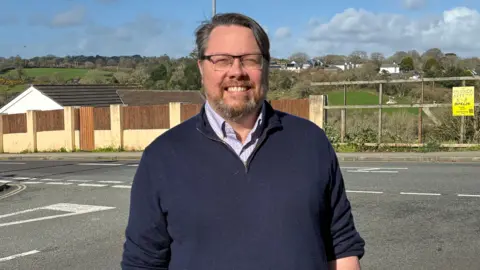 Dan has dark hair and a beard and glasses and wears a light blue shirt and navy blue quarter zip jumper. 