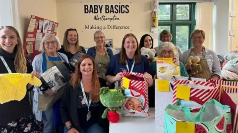 Annabel Amos/BBC A group of women of varying ages, smiling at the camera. Some are holding clothes. toys and other items. Some are holding bags full of items. The words BABY BASICS appear in blue lettering on a wall behind them.