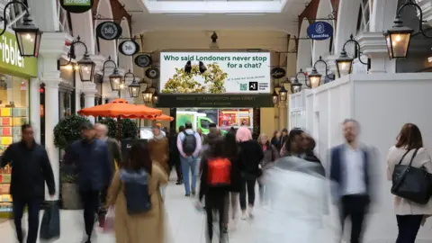 Jack McPartland/Ocean Outdoor A tall digital screen shows two starlings facing each other on a blossoming branch, with the slogan "Who said Londoners never stop for a chat?", a QR code and the London Wildlife Trust logo. Two people stop to look.