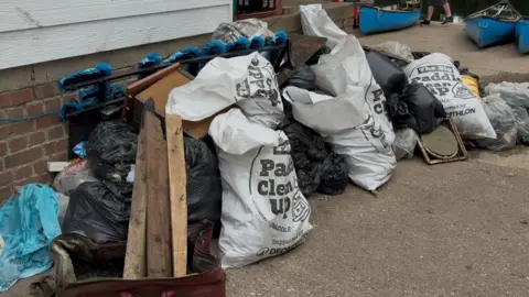BBC A pile of rubbish in bags sits at the side of a river next to three canoes.
