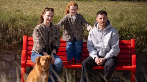 British Heart Foundation A man and woman sat on a red bench, with a young girl stood on the bench between them - a hand on each of their shoulders - and a ginger coloured dog sat in front of the woman. The woman is wearing a leopard-print brown jacket and blue jeans, is brunette and has sunglasses on top of her long hair. The man is wearing a light grey zip-up hoodie and black trousers. he has short brown hair and a moustache. The little girl is wearing a brown leopard print jacket and blue jeans. She had red hair and is wearing pink plastic frame glasses. They are all smiling and looking at the camera. Behind them is a stone wall and a patch of grass.