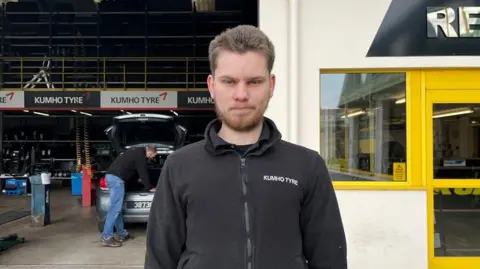 A man with short, spikey, brown hair in a black fleece standing in front of a man fixing a car in a garage. He has a tight-lipped smile.