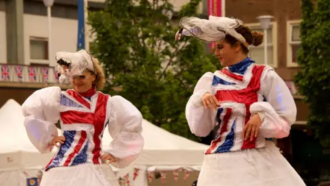 Two women on stilts with white dresses and red, white and blue sections on the front