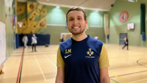 Lewis smiles at the camera as people play futsal behind him. He's wearing the blue football top with yellow sleeves. He has short brown hair and a stubbly beard.