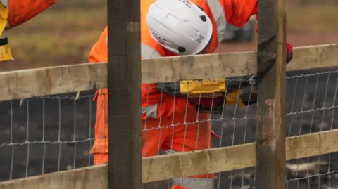 A person wearing orange high-viz and a white hard helmet is bending over holding a yellow power tool. They are working on a wooden fence. The ground is charred. 