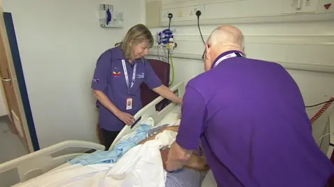 Two volunteers in purple t-shirts on either side of hospital bed of a patient whose face is obscured by the volunteer on the right