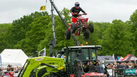 South Suffolk Agricultural Association A motorcyclist is seen flying a red quad bike over a tractor. There are crowds in the background, and behind them, green trees.