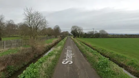 Google A google maps street view image of Sharpham Drove in Street. There is a narrow single track road, a tree and some fields in the image. There are also two stretches of water running parallel to the road. 