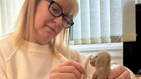 Mandy Smith Mandy Smith needle-felting a model of an animal. She is sitting at a table with her arm on the table and holding a crafting needle to sculpt the wool. She is wearing glasses and a jumper and has her head tilted concentrating as she looks at her work.