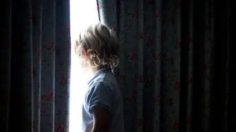 A young boy stood close to closed curtains. You cannot see his face or the room he is stood in.