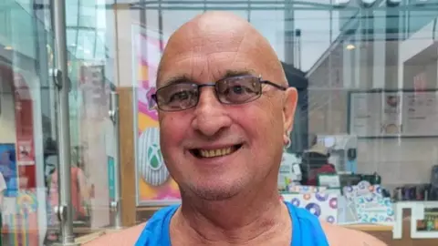 Family handout A bald man with glasses and an earring smiles at the camera. He is wearing a blue vest top.