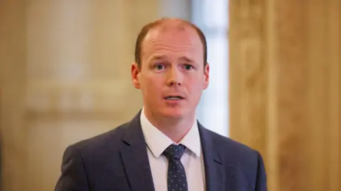 Gordon Lyons, with short brown hair, wearing a white shirt, navy tie and navy suit jacket.