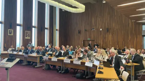 Laura Coffey/BBC Council chamber in Corby showing rows of tables set out in an arc and facing forward. Smartly dressed men and women are sat behind the desks. The desks have microphones. There is wooden panelling on the walls and two pictures.
