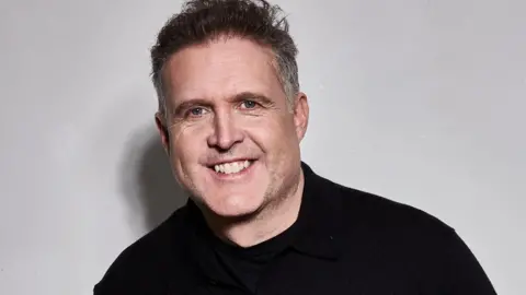 Leon John Vincent, a white man in his 50s. He is wearing a black cardigan and is standing against a grey background. He has short, brown hair, greying at the sides.