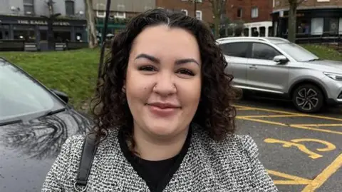Phil Harrison/BBC A woman in a grey goat with long, dark, curly hair. She is standing in a car park.
