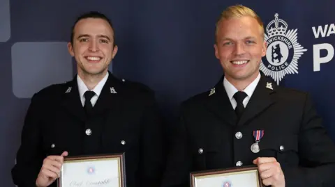 Warwickshire Police Two police officers holding certificates of commendation from the chief constable of Warwickshire Police. They are both wearing black police jackets, with a white shirt and black tie. The man on the left has short, dark hair and the man on the right has short, blonde hair with a quiff.