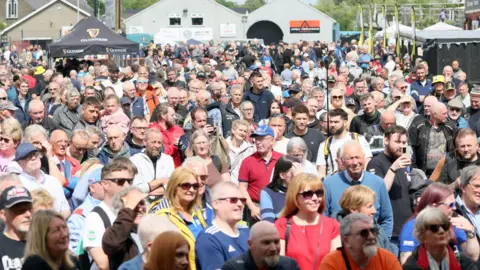 Pacemaker A large number of people standing looking towards the right hand side of the frame. Many are wearing sunglasses and hats. There are event tents in the background.