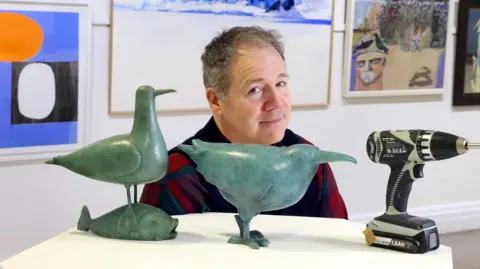 Colin Hattersley Michael Durning, a man wearing a chequed top, stands in an art gallery. In front of him is a display with several sculptures one on it, including two of birds.