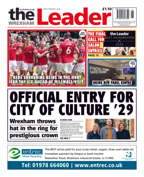 Wrexham Leader The front page of the Wrexham Leader has two women and one man standing side by side in front of a building in Wrexham city centre. Main headline: Official entry for city of culture '29'. Sub headline: Wrexham throws hat in the ring for prestigious crown. Above is a photo of the Wrexham AFC team in the team's red football kit celebrating on the pitch with the headline: Reds savouring being in the hunt for top six ahead of Millwall visit.