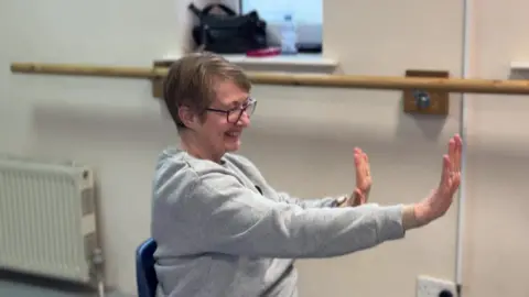 A woman sat on a chair, with her right arm extended, left arm bent with wrists flexed and fingers pointing upwards. She has a big smile on her face and is wearing a grey jumper and a pair of black rimmed glasses.