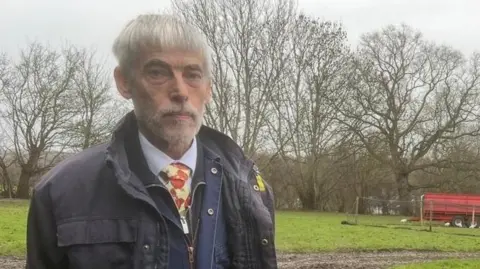 Jeff Cracknell outdoors on a misty grey day. He is wearing a dark blue wax jacket and a yellow tie with orange details. He has grey hair and dark eyebrows, and is looking at the camera with a stern expression.