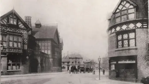 Trafford Council A black-and-white period photograph of Old Market Place in Altrincham from Trafford Council's archive. Two buildings with black-and-white, thatched-style, fronts are on either side of the road. Three horses and carts are at the end of the road.
