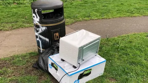 Gavin Cox A litter bin along a path in park - next to it is an abandoned black bin bag, an old printer box and microwave.