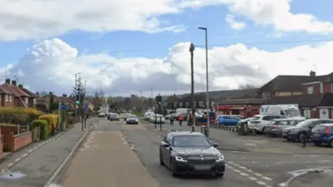 A single carriageway road, with houses on both sides, traffic lights and a junction to the right