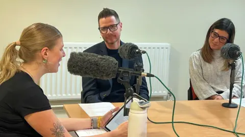 BCU Three people seated around a table recording a podcast, speaking into microphones with foam windshields; one person reads from a tablet while papers, a water bottle, and audio cables are visible on the table against a plain indoor background.
