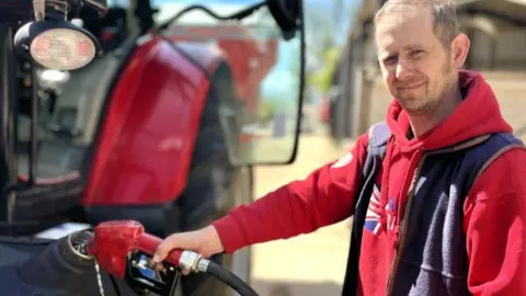 Martin Giles/BBC Philip Weston smiles at the camera as he fills his tractor with fuel. He is wearing a red-hooded jumper with a Union Jack on the front and a navy fleece gilet. His tractor is red, as is the petrol pump he is holding. 