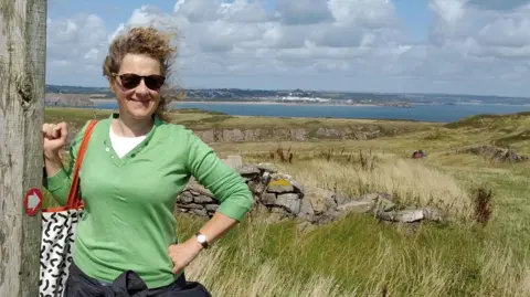 Supplied Clare Lane smiling while standing on a coastal path on a sunny day.  Clare is wearing a green sweater, white t-shirt and sunglasses. She has light brown curly hair that is blowing in the breeze.  