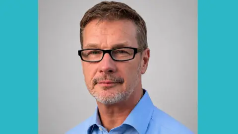 Close-up man with greying hair and greying beard, wearing glasses and a blue shirt