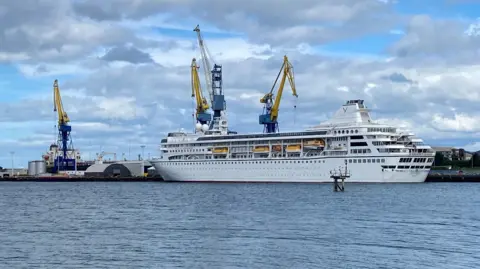 BBC A white cruise ship, docked. There are four yellow cranes and a cloudy sky in the background. 