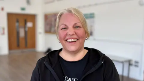 Ben Schofield/BBC Lise Gothard smiling broadly and looking down the camera. She is wearing a black, zipped hooded top, which is worn open, and a black t-shirt. She has straight hair, tied back and is also wearing two small hoop earrings. She is standing inside a large room, with a wooden floor and white walls. A set of double swing doors can be seen behind her and to the left of image.