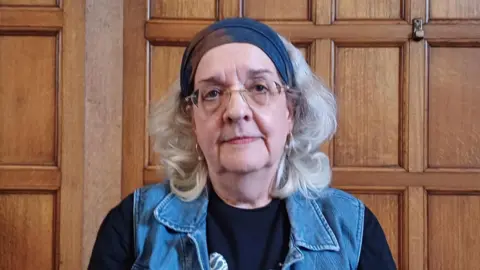 LDRS/BBC A headshot of an older woman. A polished wooden wall behind her suggests she is inside a town hall. building. She has a neutral expression and looks directly into the camera. She has grey shoulder length hair and wears glasses, hooped earrings, a navy bandana, black top and denim waistcoat.