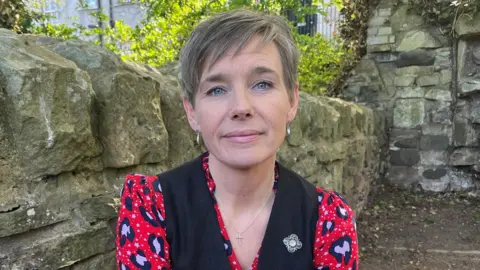 Emma Bryson, with her light brown hair in a short pixie style, looks to the camera thoughtfully against a background of a stone garden wall and greenery. She wears a red, pink and black patterned shirt with a black waistcoat and a blue and gold enamel brooch.