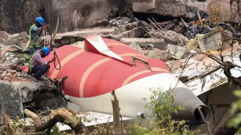 Two men wearing hard hats and shirts lower a rope at the crash site. The red and white plane is embedded into a building and surrounded by debris.