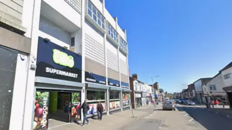 High Street in Scunthorpe taken from Google Streetview. There are people walking past a continental supermarket and cars driving down the street. It is a sunny day. 