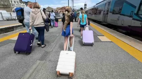 Passengers walk along a railway station platform away from the camera. They are pulling suitcases. A TransPennine Express train is stopped at the platform on one side.