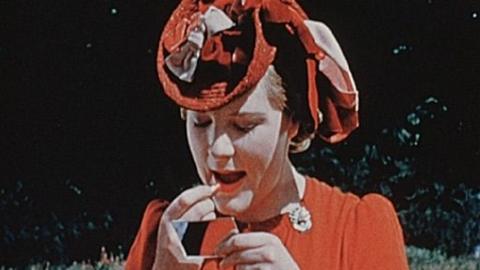 Lady in red hat and red dress, applying red lipstick whilst looking in a small hand mirror.  